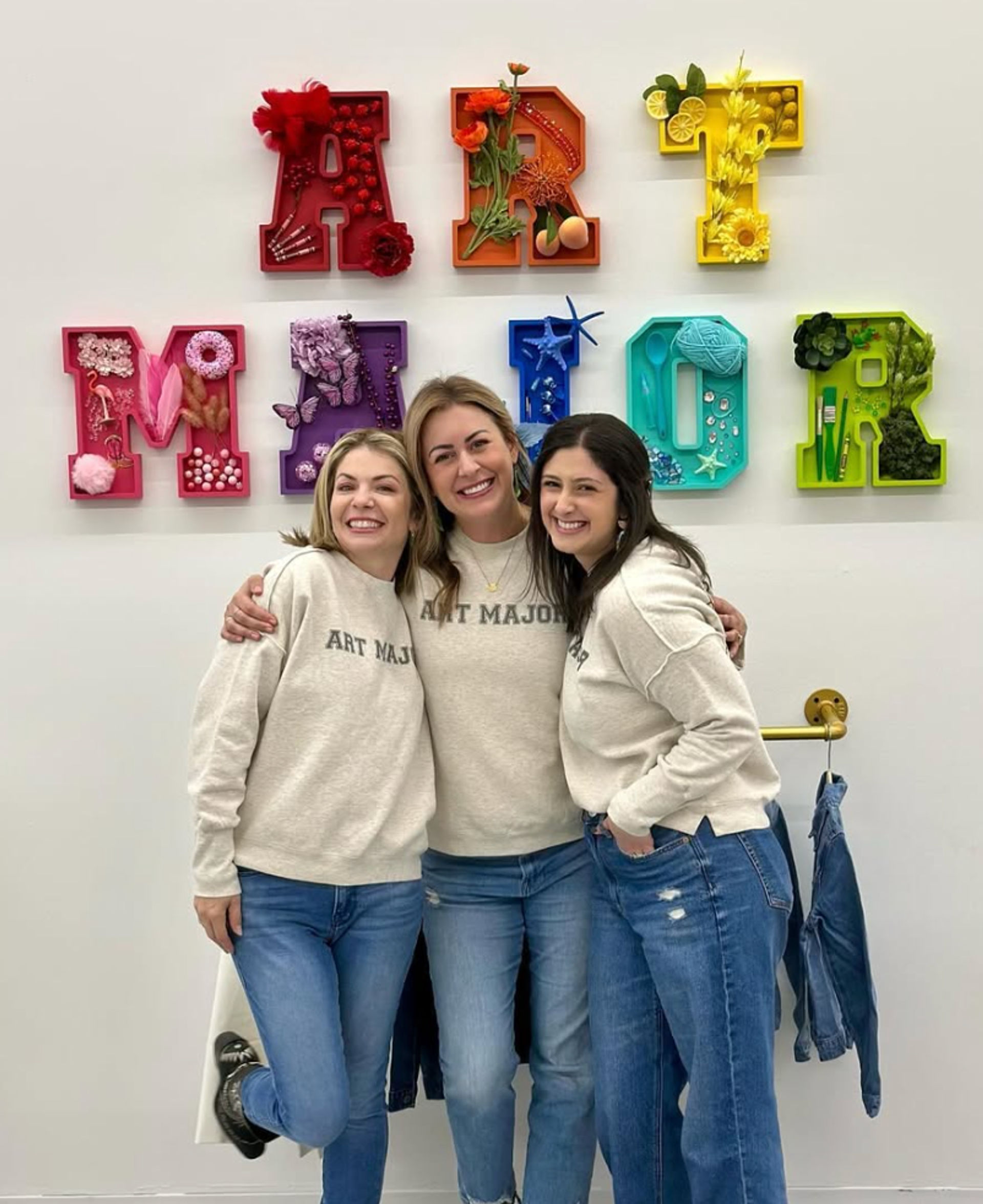 Three smiling women in matching cream "ART MAJOR" sweatshirts and jeans hugging in a bright art studio, standing in front of colorful shadow-box letters spelling "ART MAJOR" filled with craft items and flowers, with a denim jacket hanging on a nearby hook.
