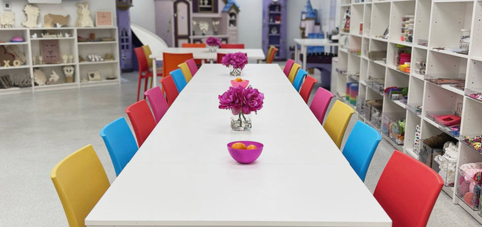 Bright indoor craft studio with a long white communal table lined with colorful red, pink, yellow and blue chairs, small vases of magenta flowers and bowls, and shelving stocked with arts-and-crafts supplies in the background.