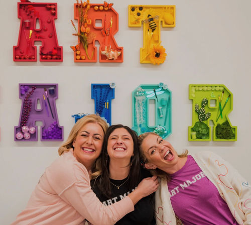 Three smiling women hugging and laughing in an art studio, posed in front of colorful shadowbox letters filled with craft supplies and decorations.