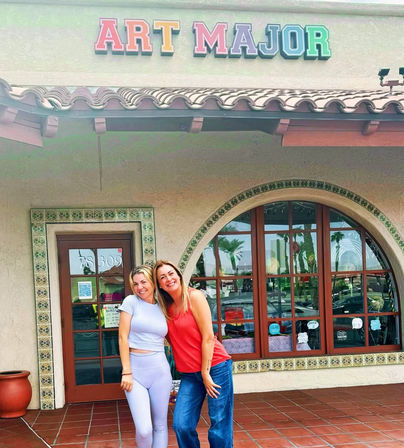 Two smiling women pose outside a Mediterranean-style art shop with stucco facade, tiled roof, arched window reflecting palm trees, and a colorful sign above.