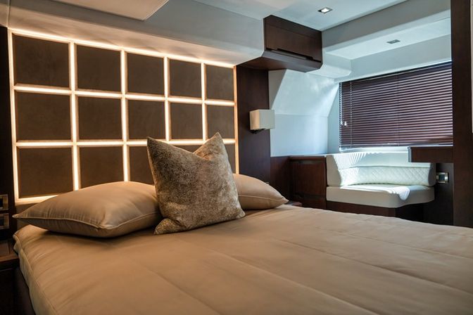 Cozy modern yacht bedroom cabin with backlit grid headboard, beige bedding and plush pillows, dark wood trim and a window seat with blinds.
