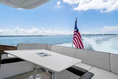 Rear deck of a cruising motorboat with white cushioned seating and table, American flag flying over the ocean wake, distant bridge and coastal skyline under a bright blue sky.