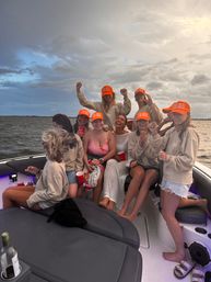 Boat party: group of friends on a motorboat on open water at dusk, wearing matching bright orange hats and neutral sweatshirts, laughing with red cups under a dramatic cloudy sky.