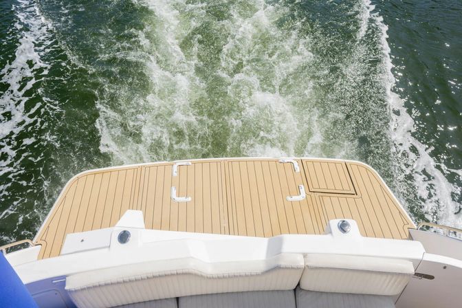 Stern view of a motorboat with a teak-style swim platform and cushioned seating, leaving white frothy wake across green water on a sunny day