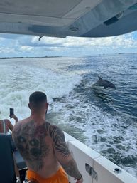 Tattooed man on a motorboat watching a dolphin surf the boat’s wake while another passenger films, with a distant bridge and cloudy sky over coastal waters.