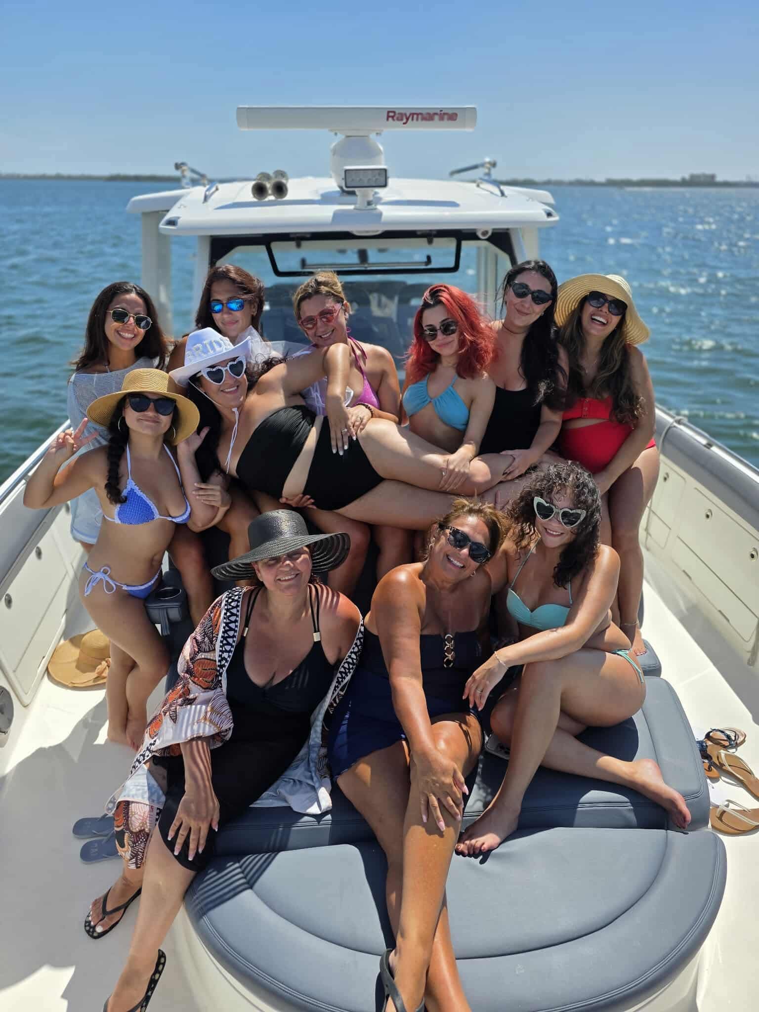 Smiling group of friends in colorful swimsuits and sun hats posing on the bow of a motorboat during a sunny coastal day with calm blue water and clear sky.