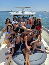 Smiling group of friends in colorful swimsuits and sun hats posing on the bow of a motorboat during a sunny coastal day with calm blue water and clear sky.