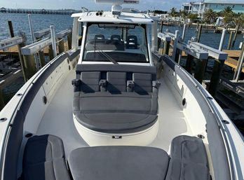 Sun-ready gray cushioned bow lounge on a white center-console boat docked at a coastal marina, showing helm cabin, cup holders, wooden docks and palm-lined waterfront in the background.