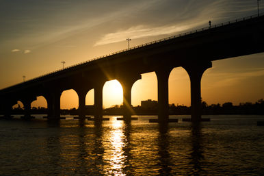 Golden sunset silhouette of an arched bridge spanning calm water, sun glowing through a central arch and reflecting on the rippling surface.