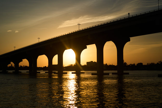 Golden sunset silhouette of an arched bridge spanning calm water, sun glowing through a central arch and reflecting on the rippling surface.