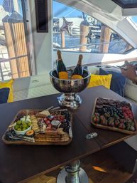 Luxury yacht interior at a marina: wooden table with two champagne bottles chilling in a silver ice bucket, a charcuterie board of cheeses, crackers, olives and berries, plus a tray of chocolate‑covered strawberries and harbor views through the windows.