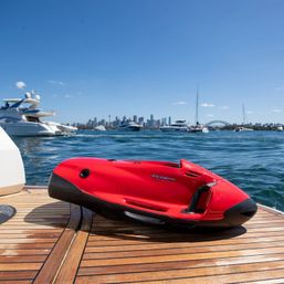 Red personal sea scooter resting on a yacht swim platform with Sydney Harbour skyline, the Harbour Bridge and yachts on sunny blue waters.