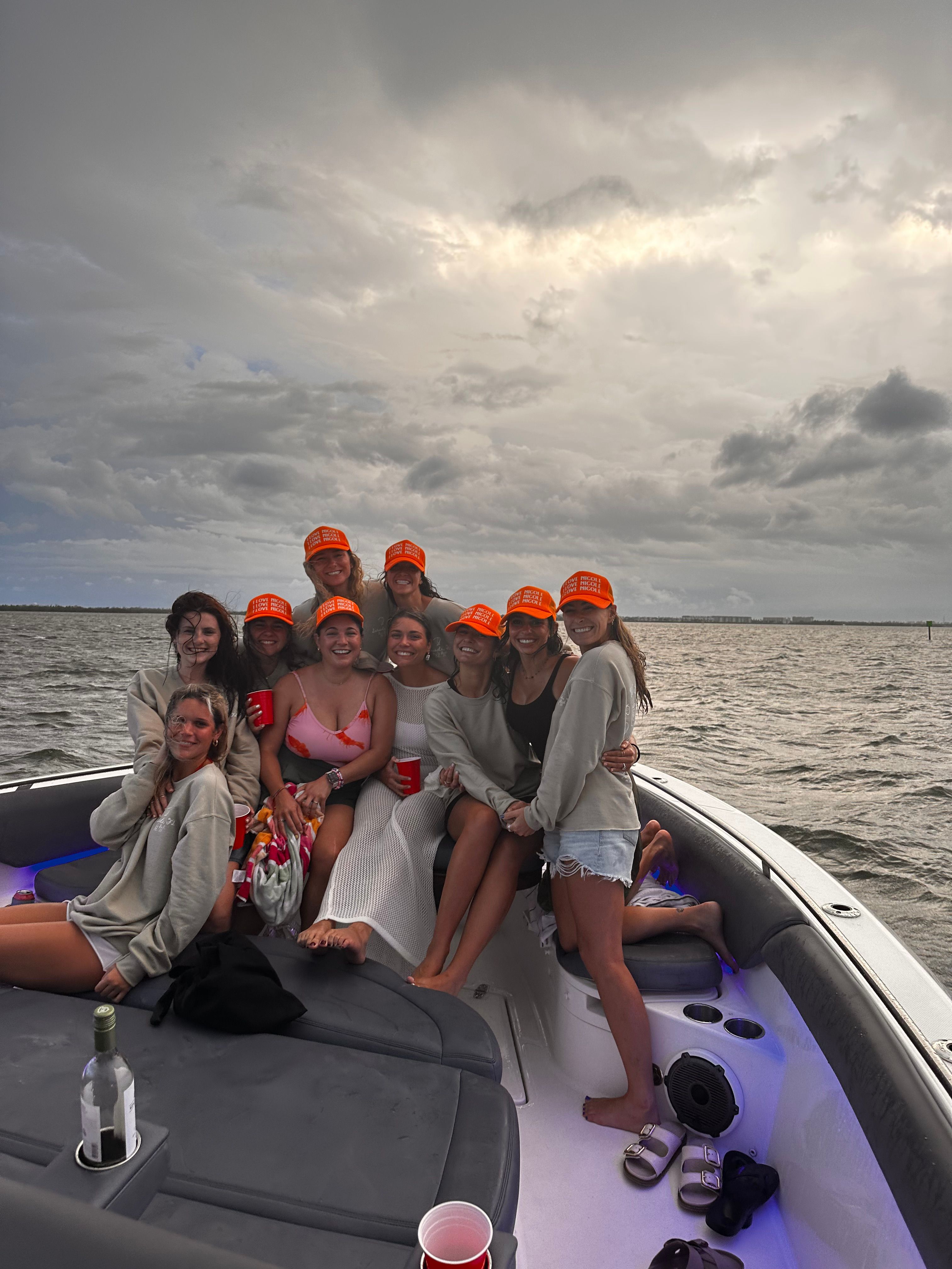 Group of friends in casual summer clothes wearing matching bright orange caps, smiling and holding red cups aboard a motorboat on coastal waters under a dramatic cloudy sky