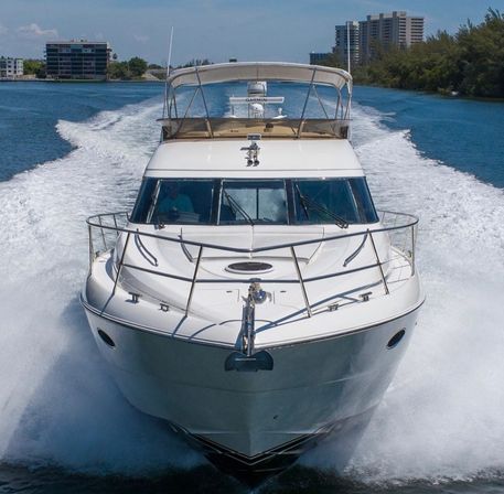 White luxury motor yacht cutting through blue coastal waterway, bow-front view with foamy wake and waterfront buildings under a sunny sky