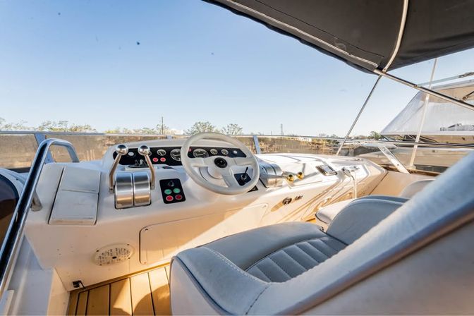 Luxury yacht helm with white steering wheel, twin throttle levers, padded captain’s seat and teak deck under a black sunshade overlooking a sunny marina
