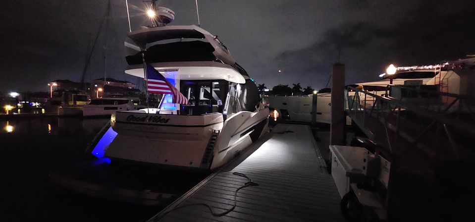 Sleek motor yacht moored at a dimly lit marina at night, glowing blue underwater lights reflecting on the water, an American flag on the stern and an illuminated dock walkway.