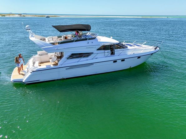 Sun-drenched white luxury motor yacht anchored in clear emerald-green coastal waters near a sandy shoreline, two people on the swim platform and an upper flybridge with black bimini and open deck seating.