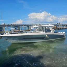 Sleek gray-and-white powerboat with four outboard engines docked at a sunny marina by a wooden pier under blue sky and puffy clouds.