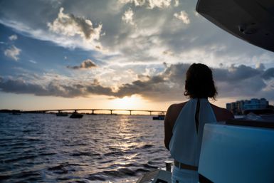 Silhouette of a person on a boat deck watching a golden sunset over a bridge, boats, and waterfront under dramatic clouds.