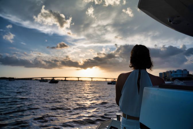 Silhouette of a person on a boat deck watching a golden sunset over a bridge, boats, and waterfront under dramatic clouds.