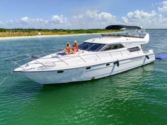 White luxury motor yacht anchored in emerald-green coastal waters near a sandy beach, three people in sun hats lounging on the bow under a bright blue sky with puffy clouds.