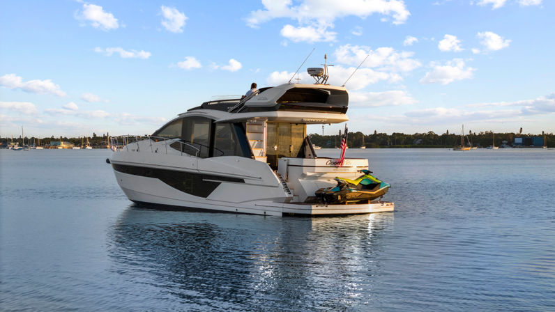 Sun-dappled white motor yacht anchored in a calm coastal marina, jet ski on the swim platform, American flag and blue sky with scattered clouds.