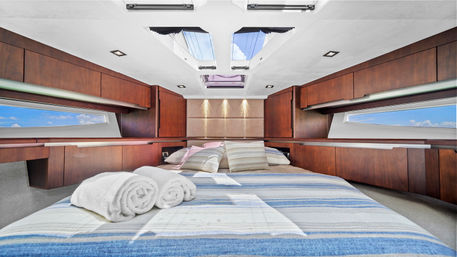 Cozy luxury yacht cabin with striped blue bedding, rolled white towels and patterned pillows, warm wood cabinetry, panoramic side windows and ceiling skylights revealing blue sky and sea views.