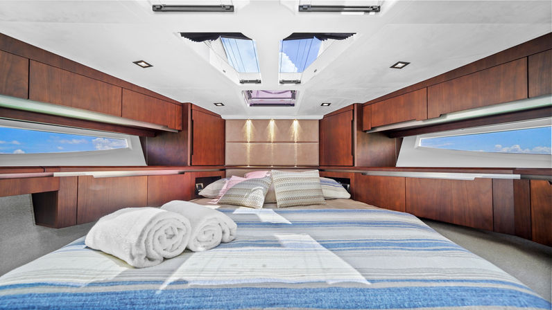 Cozy luxury yacht cabin with striped blue bedding, rolled white towels and patterned pillows, warm wood cabinetry, panoramic side windows and ceiling skylights revealing blue sky and sea views.