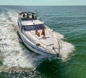 Luxury motor yacht cruising turquoise ocean on a sunny day, two people sunbathing on the bow and others on the upper deck.
