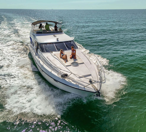 Luxury motor yacht cruising turquoise ocean on a sunny day, two people sunbathing on the bow and others on the upper deck.