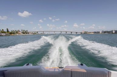 View from a motorboat stern with white wake trailing toward a low coastal bridge, turquoise water, palm-lined shore and waterfront condos under a sunny blue sky with scattered clouds.