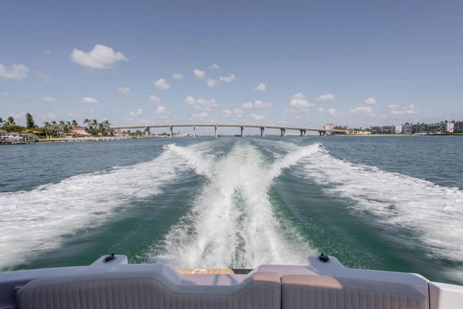 View from a motorboat stern with white wake trailing toward a low coastal bridge, turquoise water, palm-lined shore and waterfront condos under a sunny blue sky with scattered clouds.