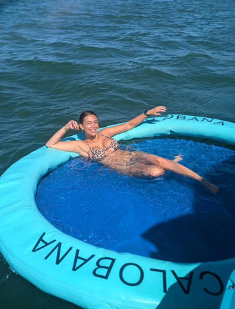 Smiling woman in a leopard-print bikini lounging with an arm draped over a turquoise circular inflatable pool float in open blue water on a sunny day.