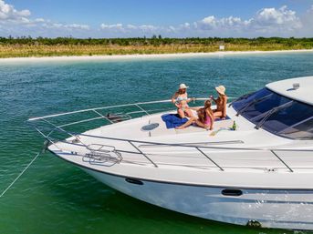 Three friends sunbathing on the bow of a white yacht anchored in green coastal waters off a sandy beach with dune grasses and a bright blue sky.
