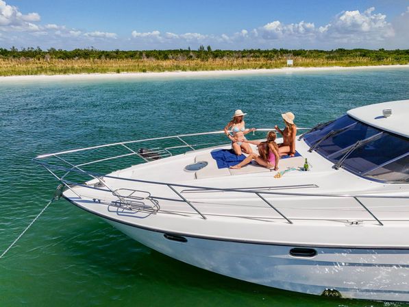 Three friends sunbathing on the bow of a white yacht anchored in green coastal waters off a sandy beach with dune grasses and a bright blue sky.
