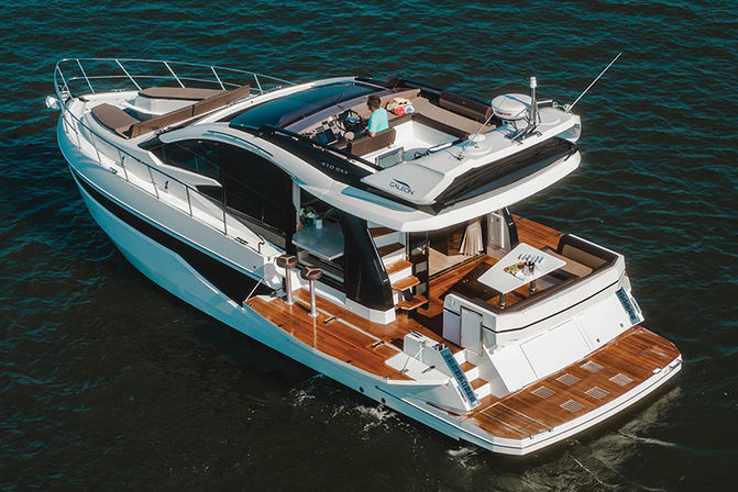 Aerial view of a sleek white luxury motor yacht on deep blue water, featuring a teak swim platform, aft cockpit dining area with seating, upper helm and sun lounges.