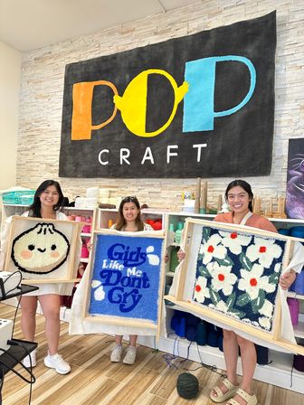 Three smiling crafters in a bright indoor craft studio holding framed tufted rug art — cute dumpling face, blue panel reading "Girls Like Me Don't Cry", and white floral pattern — yarn shelves and a large colorful craft sign on a stone wall behind them.