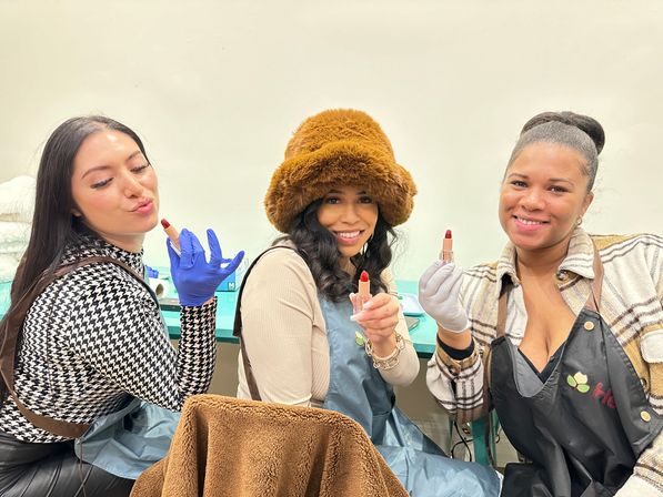 Three women in aprons and gloves at a cosmetics workshop, smiling and holding red lipsticks; middle woman wears a fuzzy brown hat