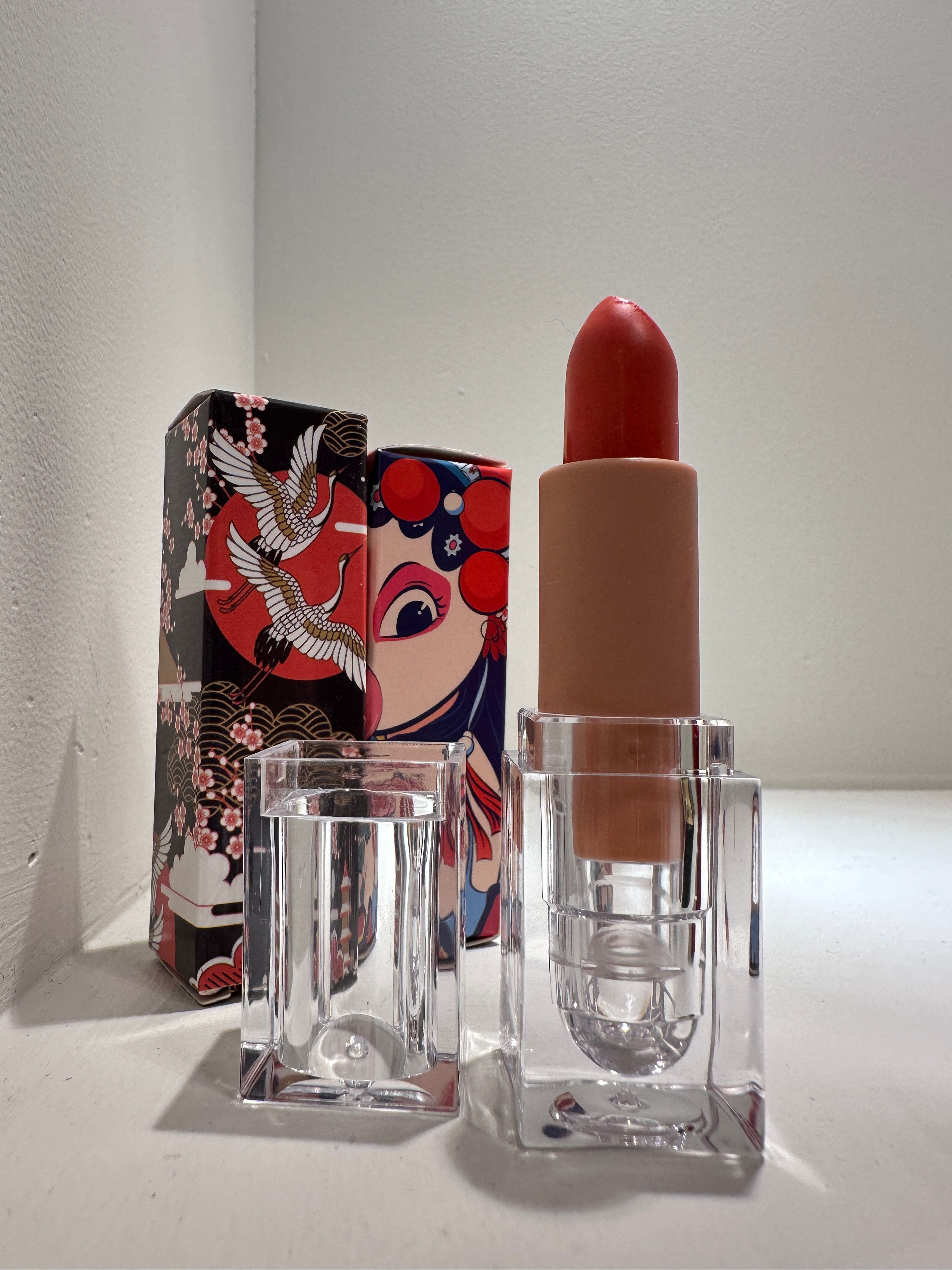 Bright red lipstick in a clear acrylic holder on a white surface, backed by two colorful Asian‑inspired decorative boxes with crane and theatrical face artwork — cosmetic packaging close-up.