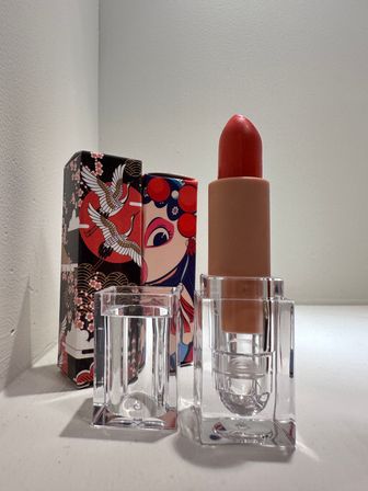 Bright red lipstick in a clear acrylic holder on a white surface, backed by two colorful Asian‑inspired decorative boxes with crane and theatrical face artwork — cosmetic packaging close-up.