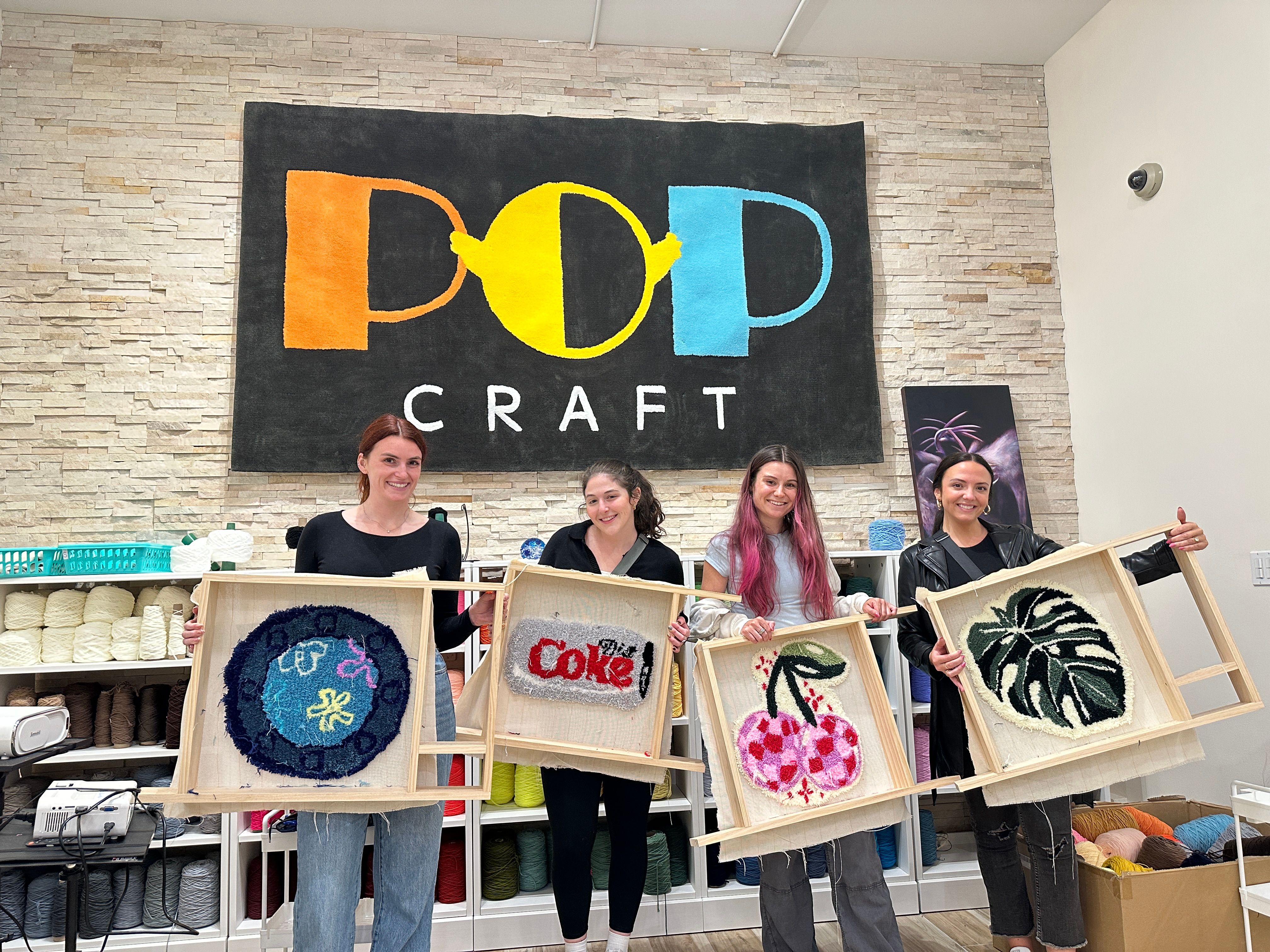 Four crafters in a bright yarn studio holding wooden frames with colorful punch-needle tufted rugs — round blue motif, red soda-style word art, pink cherries, and a green monstera leaf — yarn shelves and a large studio sign on a stone wall behind them.