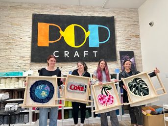Four crafters in a bright yarn studio holding wooden frames with colorful punch-needle tufted rugs — round blue motif, red soda-style word art, pink cherries, and a green monstera leaf — yarn shelves and a large studio sign on a stone wall behind them.