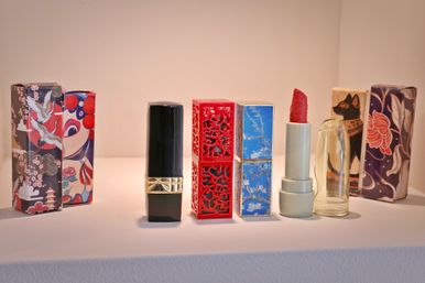 Decorative lipstick collection on a white display shelf — black tube with gold band, red laser-cut case, blue marbled case, pale green tube with sculpted red lipstick and clear cap, colorful Asian-inspired packaging behind.