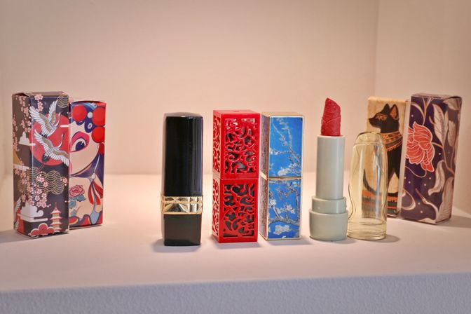 Decorative lipstick collection on a white display shelf — black tube with gold band, red laser-cut case, blue marbled case, pale green tube with sculpted red lipstick and clear cap, colorful Asian-inspired packaging behind.