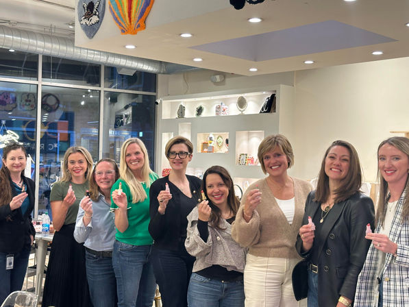Nine smiling women in a bright boutique-style shop posing together and holding small pink tubes, standing in front of lit display shelves and a storefront window.