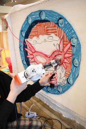 Close-up of hands with pink nail polish using an electric tufting gun to punch pink yarn into a stretched canvas, creating a colorful circular embroidery with a blue border and coral mask motif in a craft studio.