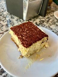 Decadent square of classic tiramisu on a white plate — creamy mascarpone layers, espresso‑soaked ladyfingers and cocoa powder dusting on a speckled stone countertop.