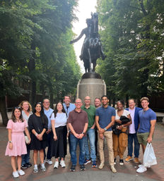 Group of people smiling and posing under leafy trees in front of a large bronze equestrian statue on a brick plaza.