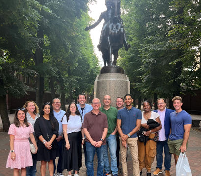 Group of people smiling and posing under leafy trees in front of a large bronze equestrian statue on a brick plaza.
