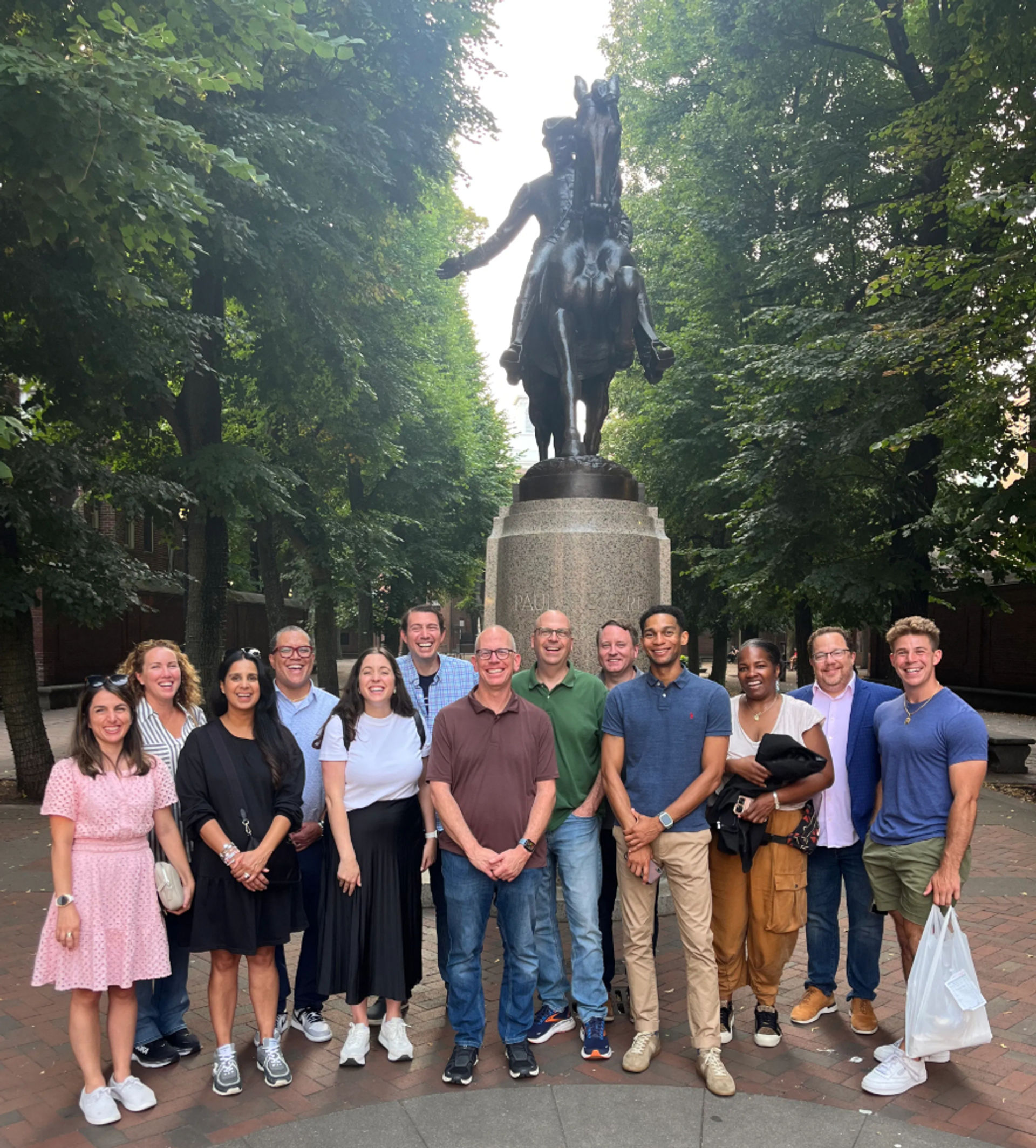Group of people smiling and posing under leafy trees in front of a large bronze equestrian statue on a brick plaza.